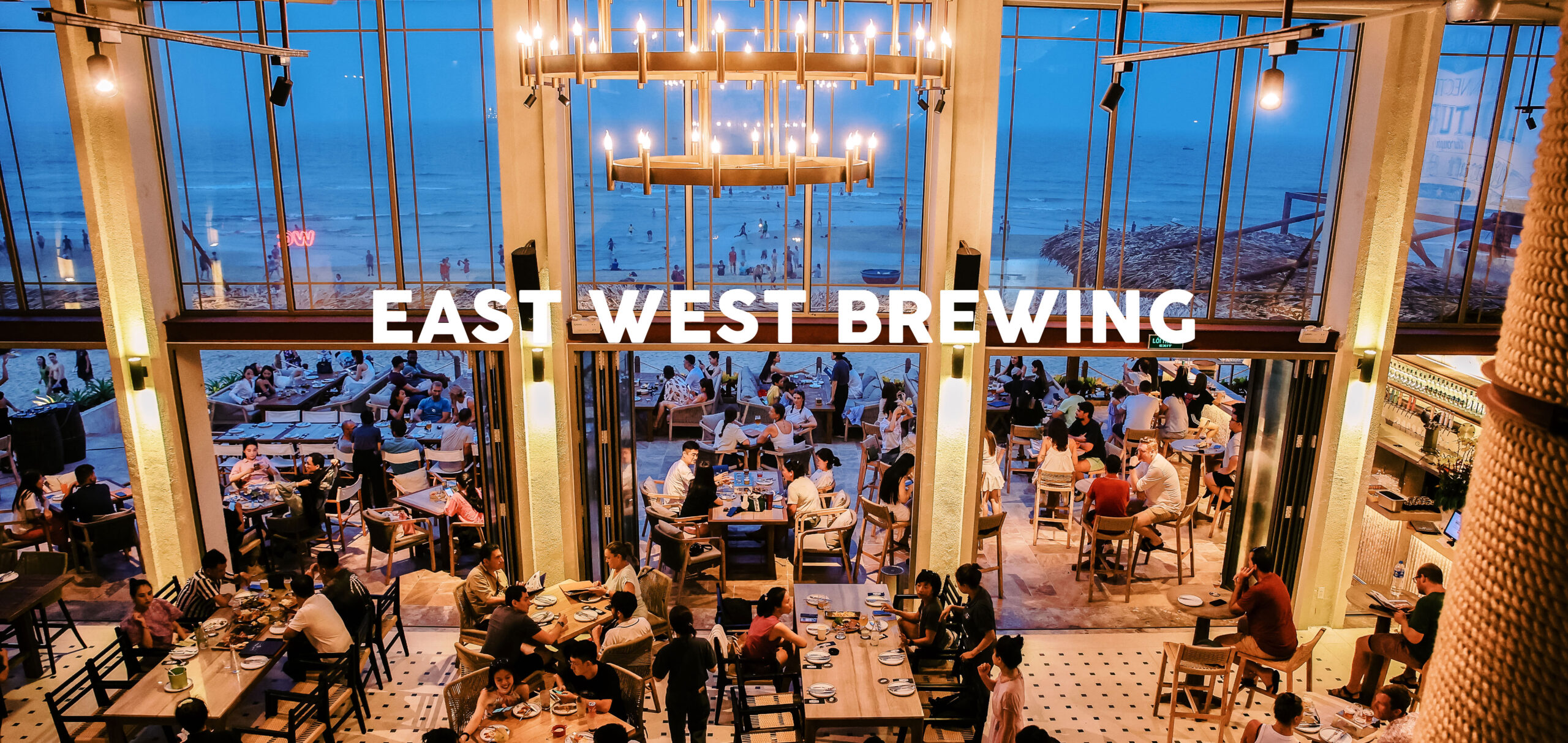 East West Brewing Da Nang interior at dusk — open-plan dining room with floor-to-ceiling glass, My Khe Beach and ocean visible behind guests