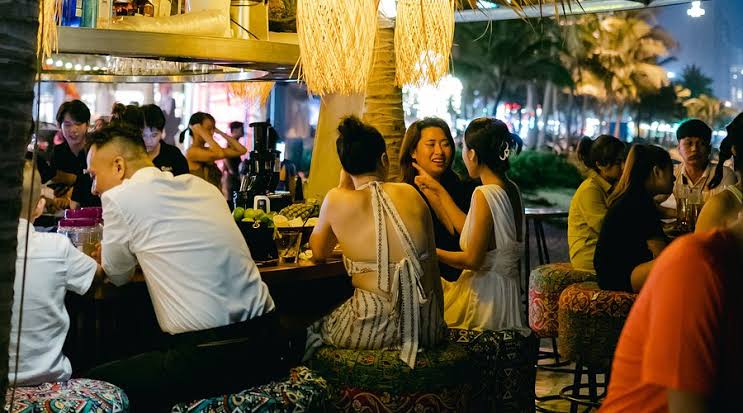 Paradise Beach Da Nang, open-air beachfront bar at night, thatched-roof terrace, guests at tables under warm lighting on My Khe