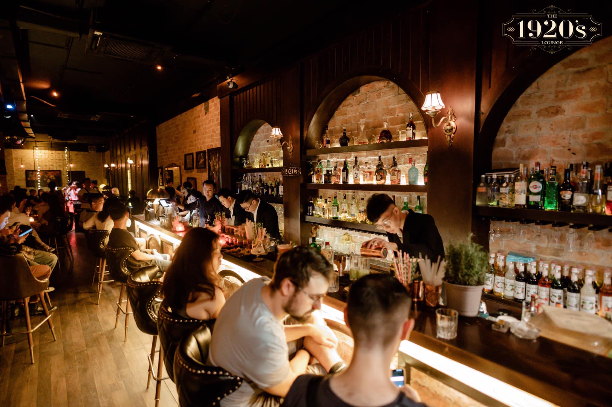 Inside The 1920's Lounge Da Nang — bartenders at the illuminated bar counter, guests seated, warm brick arches and full spirit shelves